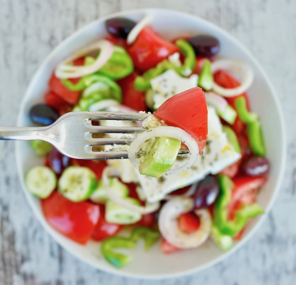 A traditional Greek salad made with tomatoes, cucumbers, olives, pepper, onion and feta in a white bowl.